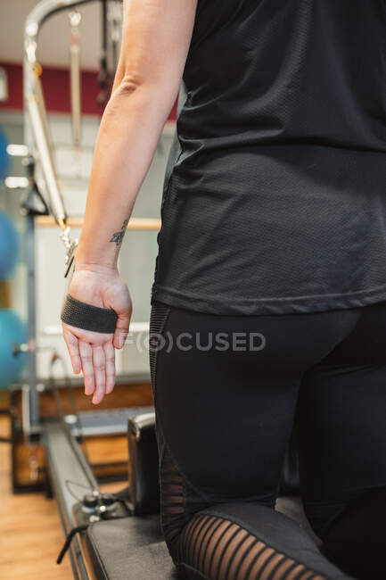 Vista posteriore della femmina adulta in abbigliamento sportivo seduta sulla macchina pilates e facendo esercizi con bande di resistenza — Foto stock