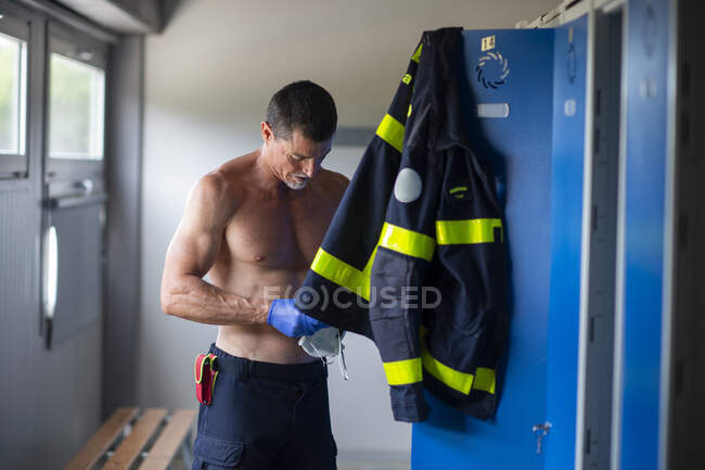 Bombero serio con el torso desnudo y en guantes de látex de pie con mascarilla respiradora cerca del armario en la estación de bomberos mientras se prepara para el trabajo - foto de stock