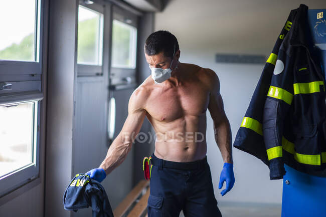 Pompier sérieux avec torse nu et gants en latex debout avec masque respiratoire près du casier à la caserne de pompiers tout en se préparant pour le travail — Photo de stock