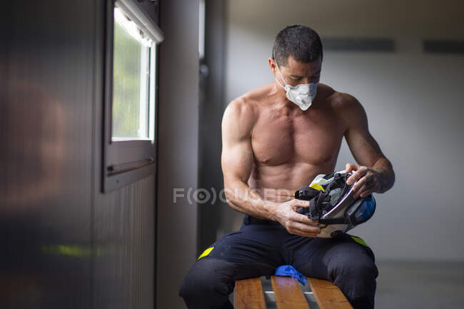 Pompier mâle mature fort avec torse nu assis sur le banc dans le masque et tenant un casque tout en regardant loin — Photo de stock