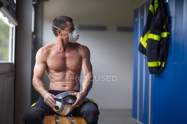 Сильный взрослый пожарный с обнаженным туловищем, сидящий на скамейке в маске и держащий шлем, отводя взгляд — стоковое фото