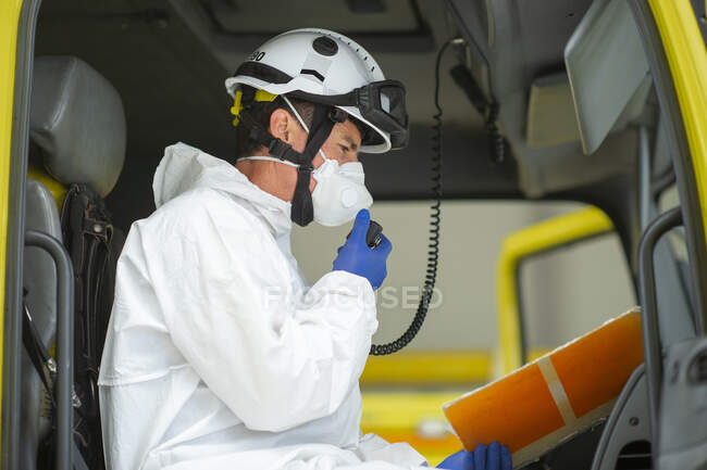 Vista lateral del bombero con uniforme protector y casco duro con guantes médicos sentados en el camión de bomberos hablando en walkie talkie - foto de stock