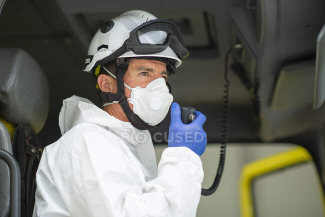 Bombero vista lateral con respirador y casco sentado en coche de bomberos y usando walkie talkie - foto de stock