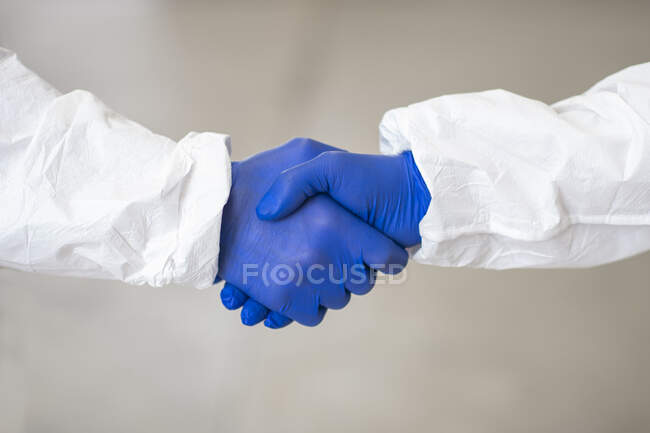 Vista lateral de practicantes irreconocibles de coworking con disfraces y guantes médicos estrechando las manos durante la epidemia de coronavirus - foto de stock