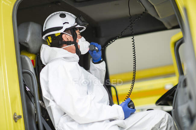 Bombero vista lateral con respirador y casco sentado en coche de bomberos y usando walkie talkie - foto de stock