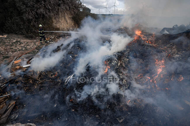 Side view of brave fireman in protective uniform standing with hose and extinguishing fire on dump in mountains — Stock Photo