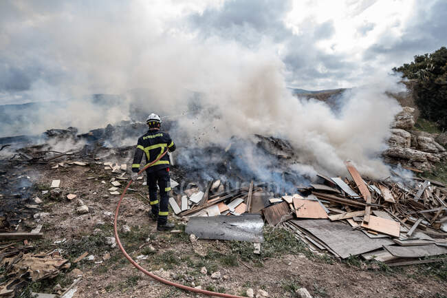 Rückansicht des tapferen Feuerwehrmannes in Schutzuniform, der mit Schlauch steht und Feuer auf Müllkippe in den Bergen löscht — Stockfoto