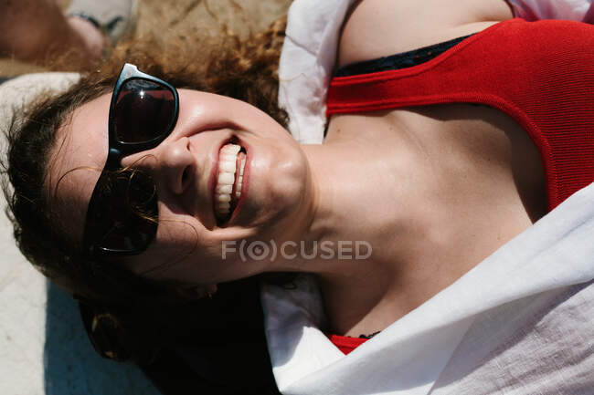 Vista superior de una mujer sonriente con gafas de sol y atuendo de verano tumbada en la costa pedregosa y disfrutando del fin de semana - foto de stock