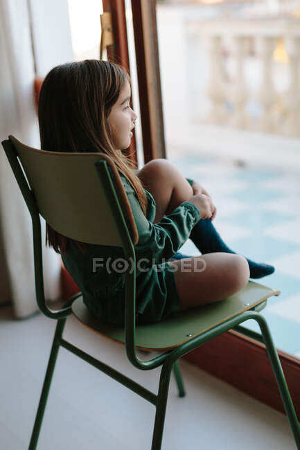 Вид збоку вдумливої дитини, що розслабляється на стільці і спостерігає за вулицею, дивлячись з вікна — стокове фото