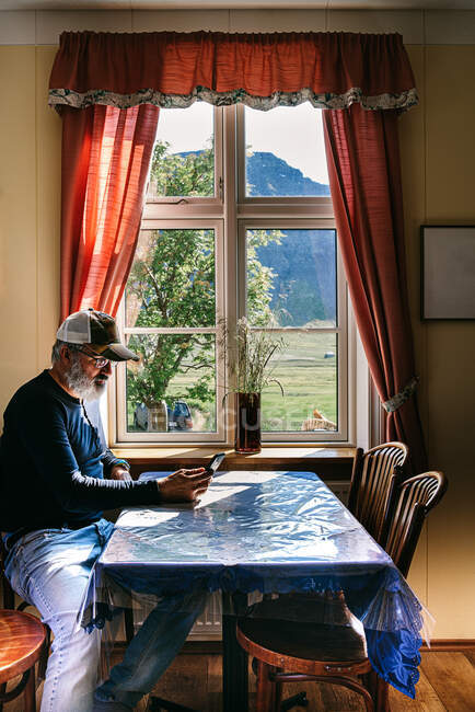 Vista lateral de anciano barbudo hombre sentado en la mesa en el acogedor comedor cerca de la ventana y leer mensajes en el teléfono móvil mientras se relaja durante el fin de semana - foto de stock