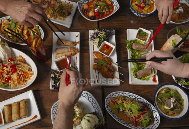 Вид сверху на пряную тайскую еду, подаваемую на деревянном столе, руки людей с палочками — стоковое фото