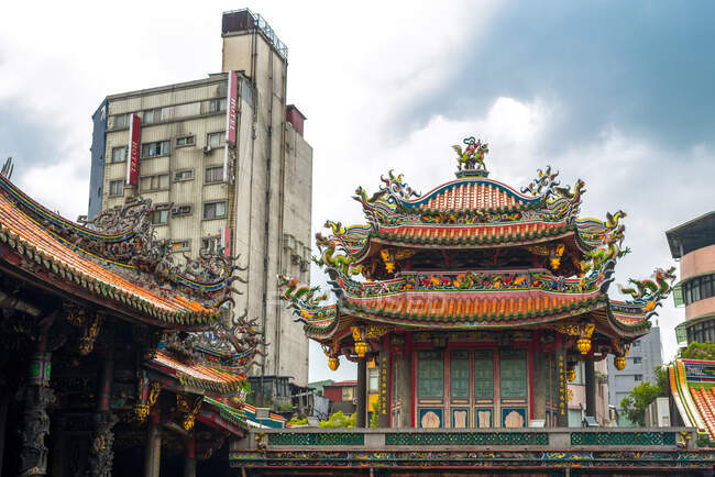 Angolo basso del tradizionale tempio Bangka Lungshan con design ornamentale situato vicino all'edificio moderno della città contro il cielo nuvoloso a Taiwan — Foto stock