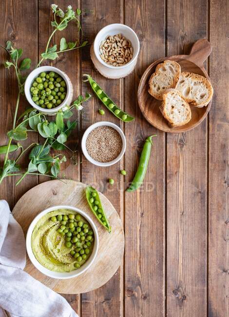 Top view composition with bowl with hummus made with green pea arranged on wooden table with ingredients for recipe and bread slices — Stock Photo