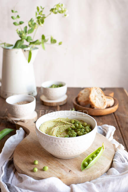 Composición con cuenco con hummus hecho con guisante verde arreglado en mesa de madera con ingredientes para la receta y rebanadas de pan - foto de stock