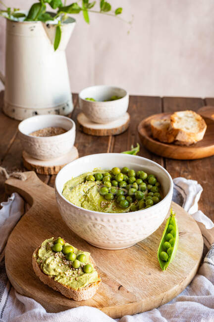 Composizione con ciotola con hummus a base di piselli verdi disposti su tavola di legno con ingredienti per la ricetta e fette di pane — Foto stock