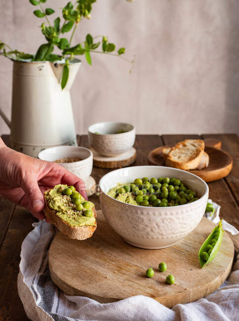 Mão segurando saboroso brinde saudável com hummus ervilha verde caseiro durante o café da manhã em casa — Fotografia de Stock