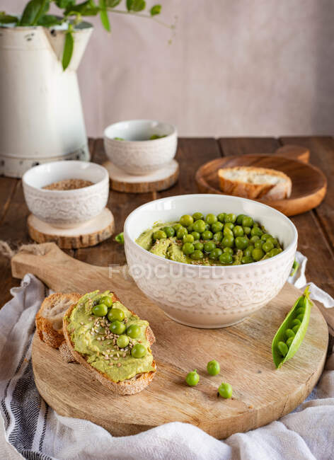 Композиція з мискою з гумусом, зроблена з зеленого гороху, розташованого на дерев'яному столі з інгредієнтами для рецепту та скибочками хліба — стокове фото