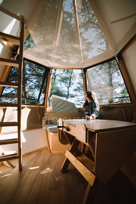 Feminino em desgaste casual sentado à mesa em casa de madeira futurista localizado no acampamento na floresta no dia ensolarado — Fotografia de Stock