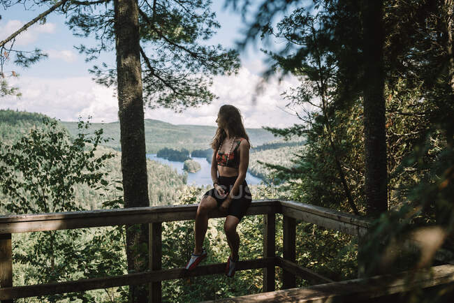 Full body of young female tourist sitting on railing of wooden terrace and looking away in forest of La Mauricie National Park in Quebec, Canada — Stock Photo