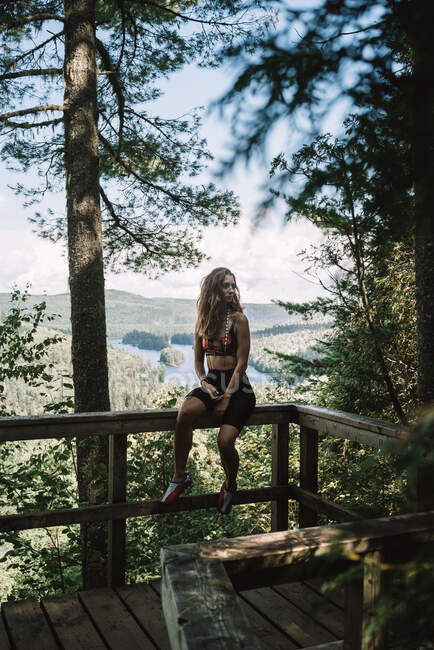 Full body of young female tourist sitting on railing of wooden terrace and looking away in forest of La Mauricie National Park in Quebec, Canada — Stock Photo