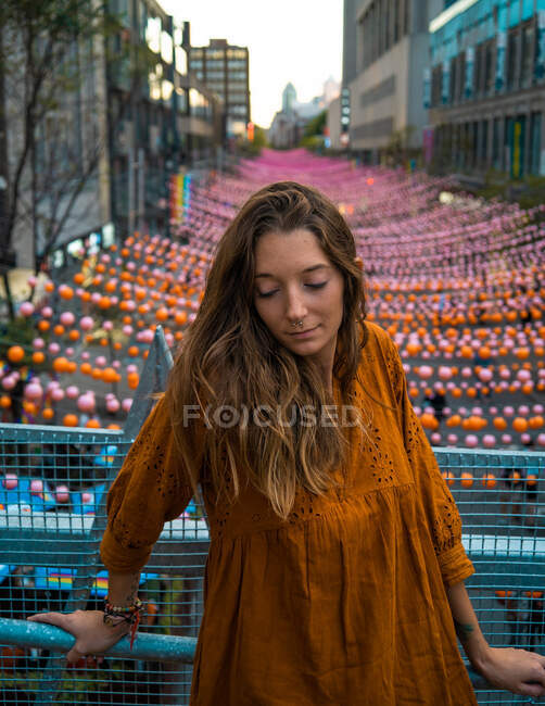 Calm brunette in dark orange dress standing on bridge with view of Montreal street decorated with multicolored garlands with eyes closed — Stock Photo