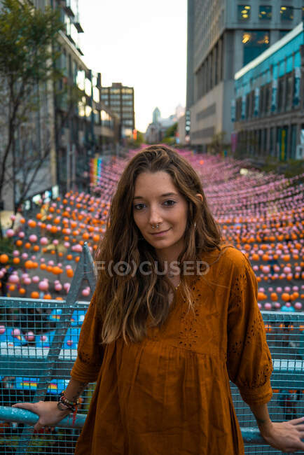 Ruhige Brünette in dunkelorangefarbenem Kleid steht auf einer Brücke mit Blick auf die Straße von Montreal, dekoriert mit bunten Girlanden und blickt in die Kamera — Stockfoto