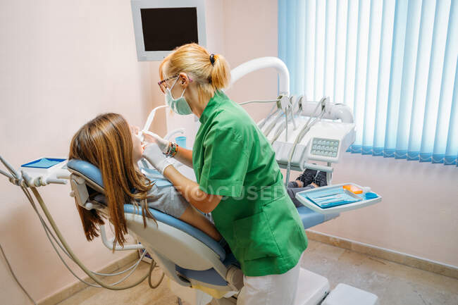 Профессиональный врач средних лет в зеленой форме осматривает полость рта женщины в стоматологическом кресле — стоковое фото