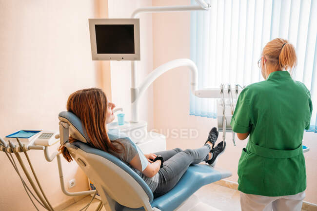 Médico profesional de mediana edad enfocado en uniforme verde que examina la cavidad oral de la mujer en la silla del dentista - foto de stock