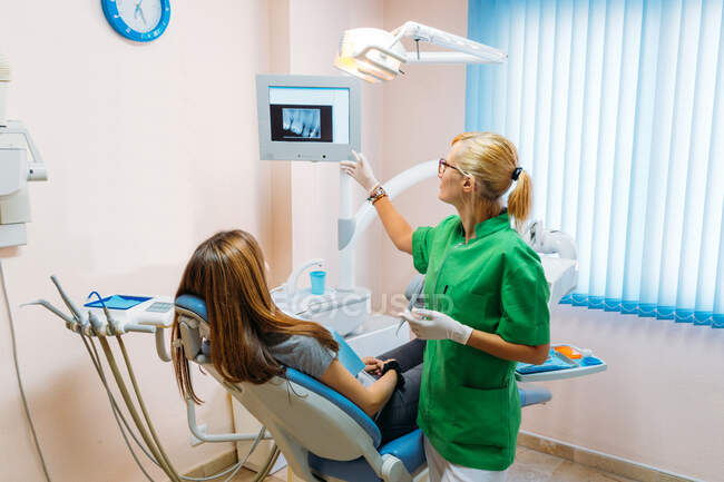Donna adulta in guanti medici e uniforme che spiega il risultato dei denti ai raggi X al paziente in ufficio — Foto stock