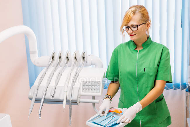 Dentista femenina adulta en uniforme verde y gafas que preparan los instrumentos para la visita del paciente en la oficina del hospital - foto de stock