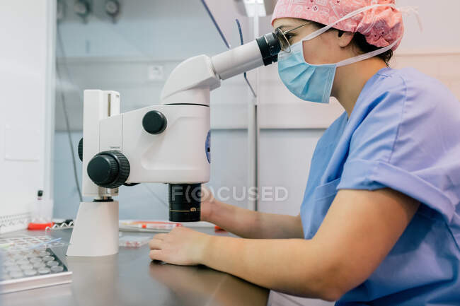 Vista lateral de la mujer en uniforme médico y máscara con microscopio moderno para examinar las células humanas mientras trabaja en el laboratorio de la clínica moderna - foto de stock