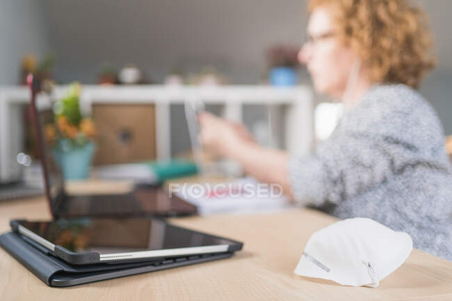 Side view of adult woman in casual clothes and eyeglasses using smartphone in earphones while sitting at table and working on laptop at home — Stock Photo