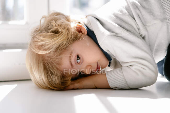 Seitenansicht eines ruhigen blonden Jungen in Freizeitkleidung, der an sonnigen Tagen auf der Fensterbank in einer modernen Wohnung in die Kamera blickt — Stockfoto