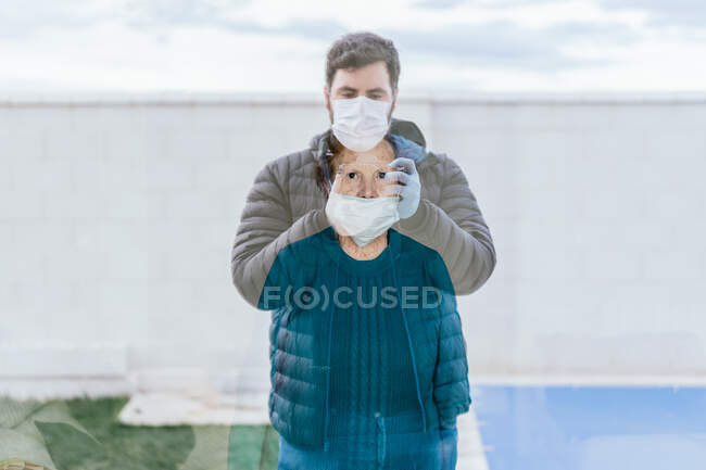 Dark haired young male in warm jacket and medical mask taking photo of senior woman in medical mask through window while standing outside in front of modern hospital — Stock Photo