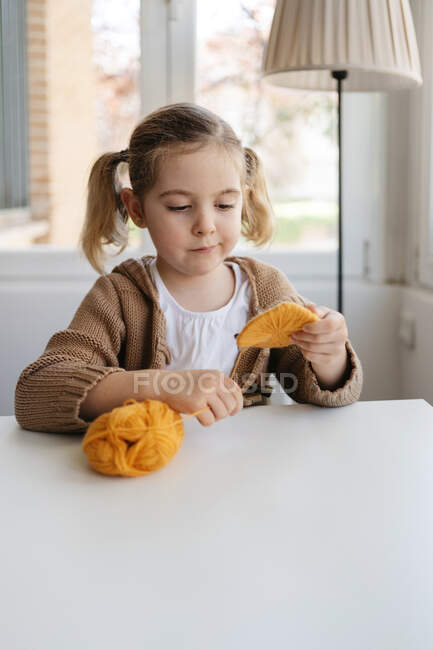 Маленькая девочка в повседневной одежде обмотки шерстяные нити на картон шаблон, чтобы сделать ручной помпон для шляпы в уютной гостиной — стоковое фото