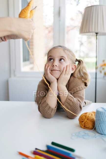 Cute blond girl in warm sweater leaning on hand and looking at babysitter showing craft of woolen thread in modern kindergarten — Stock Photo