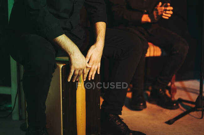 Crop anonymous male artists in black wear sitting on chairs and playing Peruvian musical instrument cajon on wooden platform in auditorium - foto de stock