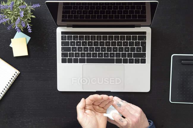 From above crop male treating hands with sanitizer for protection from coronavirus while working on laptop at home during quarantine — Stock Photo