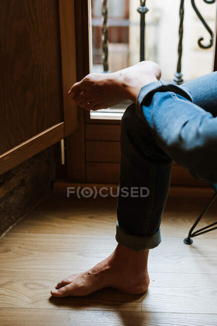 Unrecognizable barefoot person in jeans sitting near window and relaxing in cozy room at home — Stock Photo