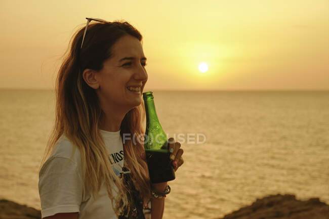 Smiling young lady with bottle of beer during sunset on seashore — Stock Photo