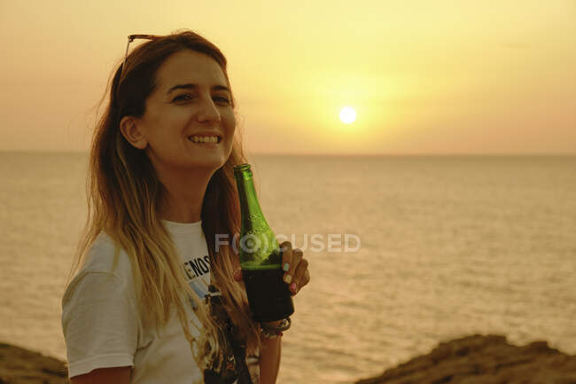 Feliz joven viajera en ropa casual y gafas de sol sosteniendo botella de cerveza de vidrio mientras se relaja al atardecer en la costa y mirando a la cámara en Ibiza - foto de stock