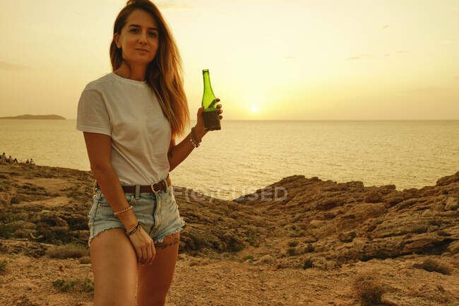 Jovencita sonriente con botella de cerveza al atardecer en la orilla del mar - foto de stock