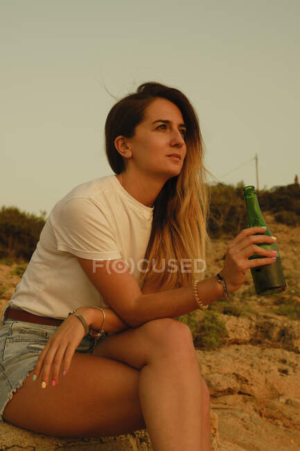 Young lady with bottle of beer during sunset on seashore — Stock Photo