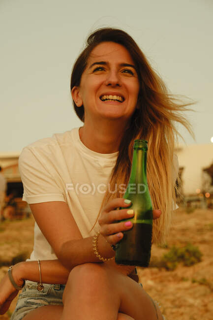 Усміхнена молода леді з пляшкою пива під час заходу сонця на березі моря. — стокове фото