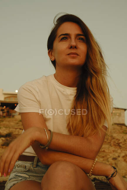Low angle of woman in casual clothes sitting on rocky seashore and looking away dreamily during sunset in Ibiza — Stock Photo