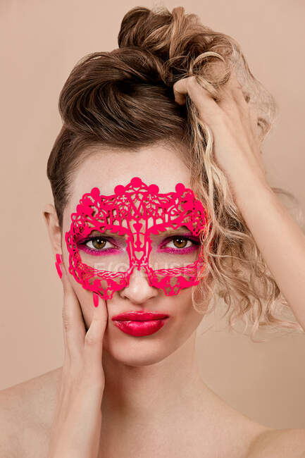 Junge Frau mit lebhaftem Make-up und ornamentaler Maske, die Gesicht und Haare vor beigem Hintergrund berührt — Stockfoto