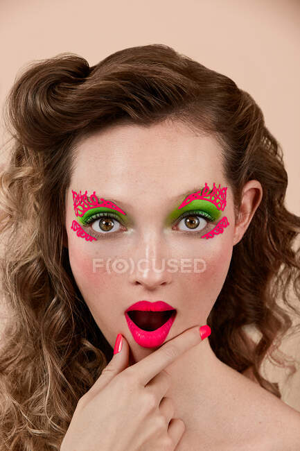 Jeune femme étonnante avec un maquillage coloré frottant le menton et regardant la caméra avec la bouche ouverte sur fond beige — Photo de stock