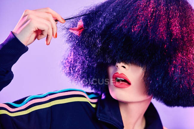 Donna stupita in elegante abbigliamento sportivo ispezionare i capelli finti sotto illuminazione brillante su sfondo viola — Foto stock