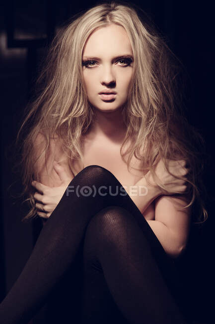 Joven modelo femenina rubia en medias que cubren el pecho desnudo y mirando a la cámara en habitación oscura - foto de stock