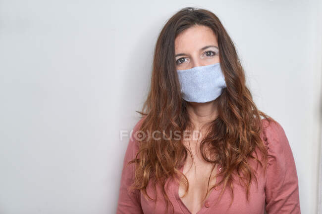 Erwachsenes Weibchen in perlrosa Bluse mit langen braunen Haaren und blauer hausgemachter Maske steht neben weißer Wand und blickt während der Pandemie in die Kamera — Stockfoto